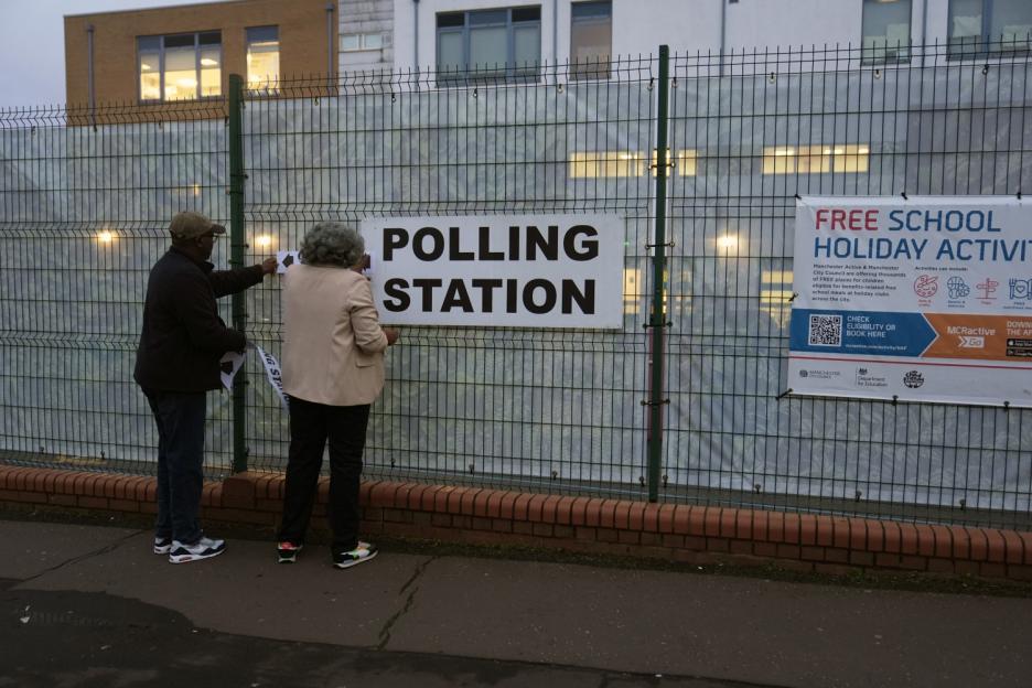 Two people attach a "Polling Station" sign to a fence.