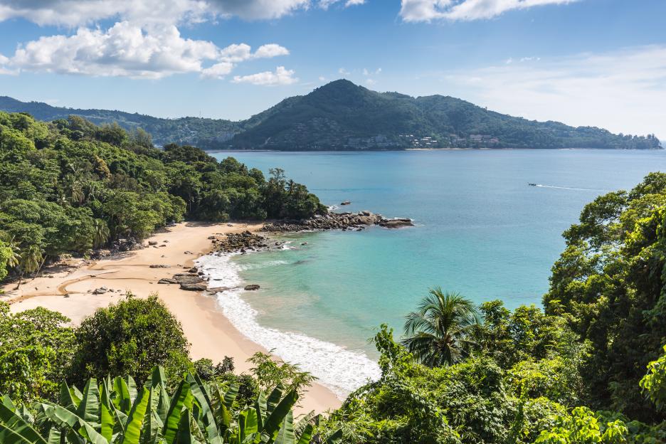 Top view of a tropical coastline in Phuket, Thailand, with a sandy beach, turquoise water, lush green rainforest, and distant mountains.