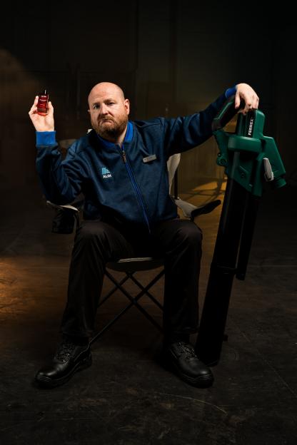 A man in an Aldi uniform sits in a chair, holding a bottle of perfume and a leaf blower.