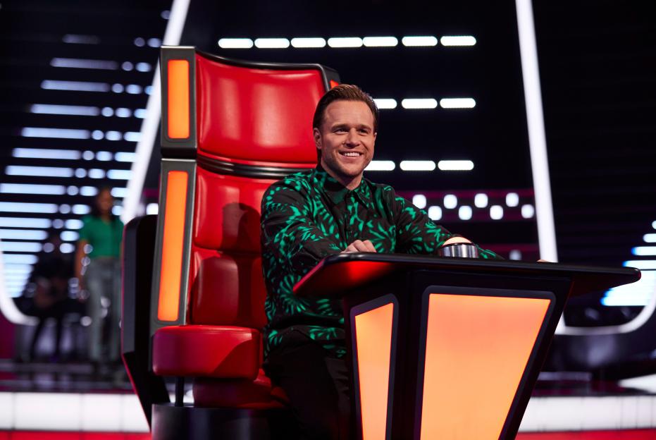 Olly Murs smiling in a red coach's chair on "The Voice UK."