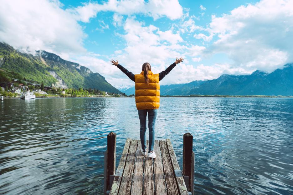 Young woman with arms outstretched on a wooden bridge, enjoying the view of Lake Geneva and mountains in Switzerland.