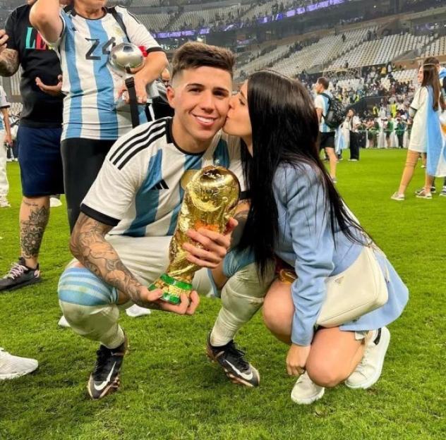 Football player Enzo Fernandez with the World Cup trophy, being kissed on the cheek by a woman.