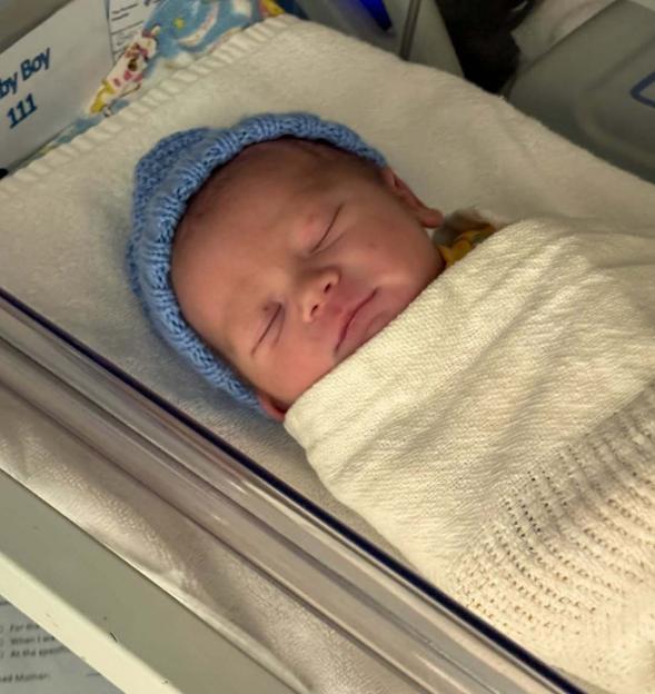Baby Bowden Mark Richard Sugg in a blue knitted hat, wrapped in a white blanket.