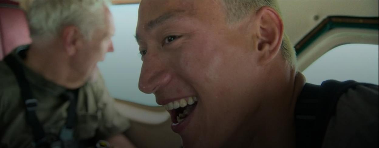 A man with short blonde hair laughing next to another man in a vehicle.