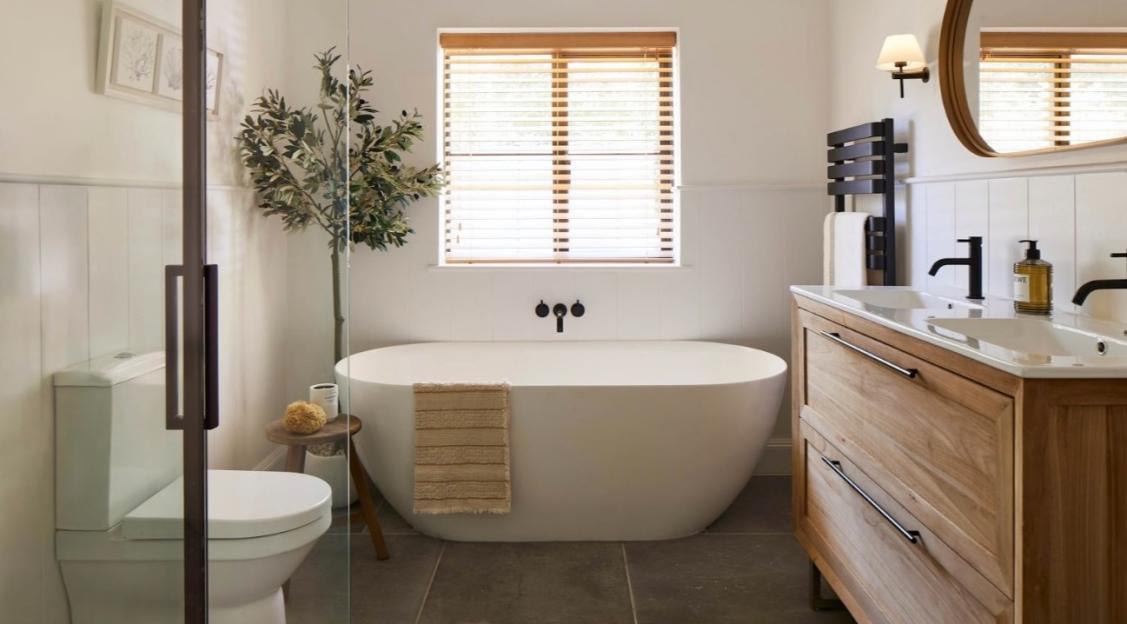 A spacious bathroom with a modern freestanding bathtub, a toilet in a glass enclosure, a wooden double vanity with two sinks, and a large window with wooden blinds.