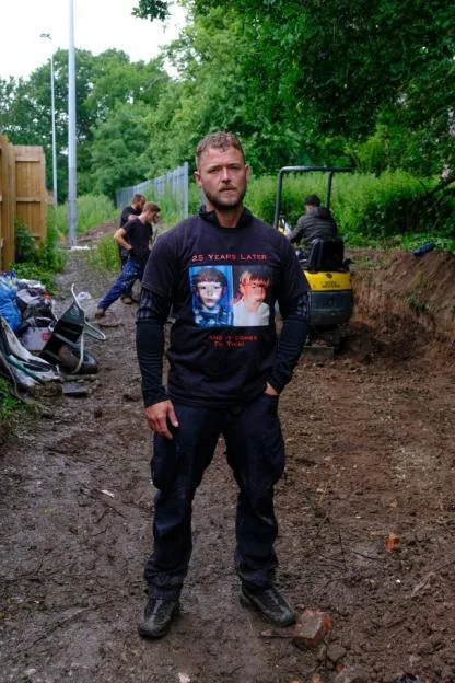A man standing on a muddy path wearing a black t-shirt that says "25 years later and it comes to this!" with two childhood photos printed on it.