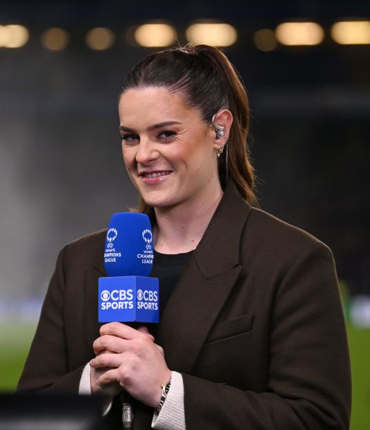 A smiling female commentator wearing a dark brown jacket holds a CBS Sports microphone on a blurred sports field.