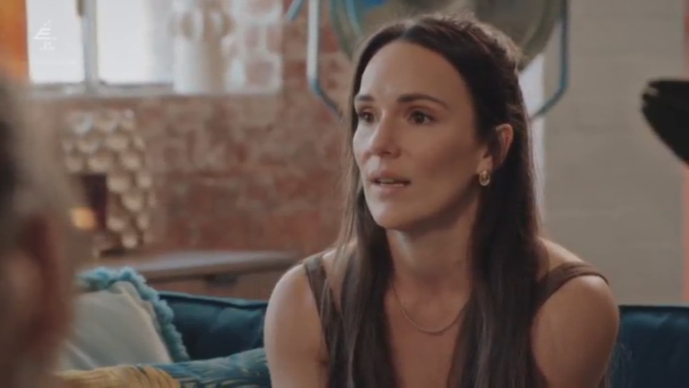 A woman speaking, with brown hair and wearing a brown top and gold necklace.