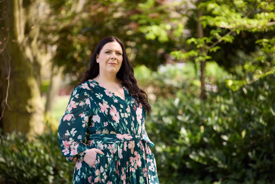 A woman in a floral dress with long dark hair poses in a park.