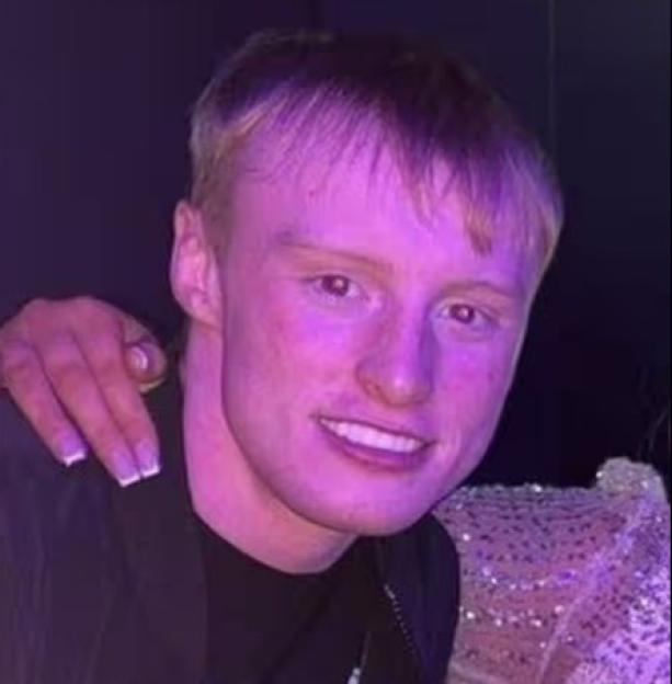 A young man with light hair and a black jacket smiles at the camera, with a woman's hand on his shoulder.