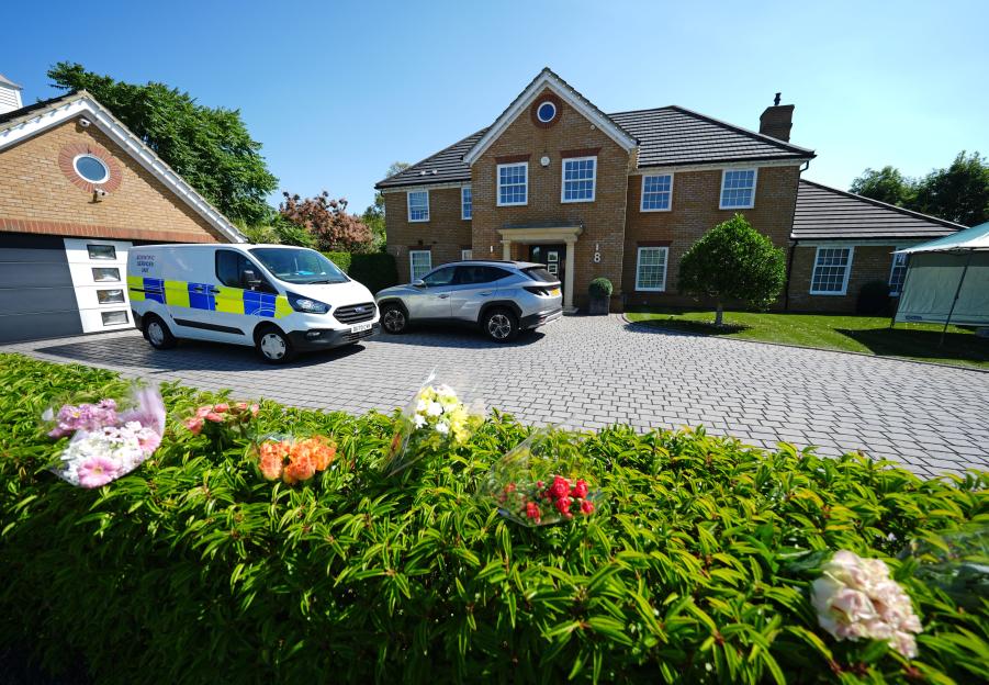 A "Scientific Services Unit" van and a car are parked in the driveway of a two-story brick house, with bouquets of flowers laid on a hedge in the foreground.