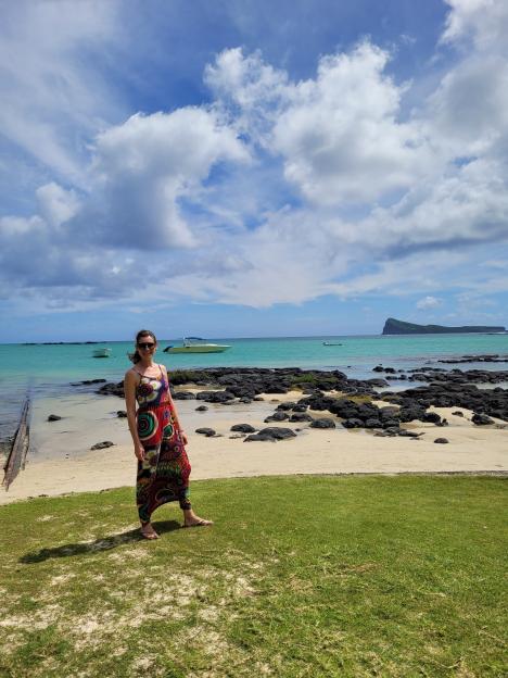 Michele Akester-Marsh on a beach in Mauritius, wearing a colorful patterned jumpsuit and sunglasses, with boats and an island in the background.