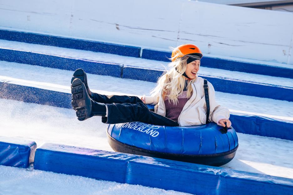 A young woman with blonde hair, wearing a white jacket and an orange helmet, smiles while snow tubing down a slope.