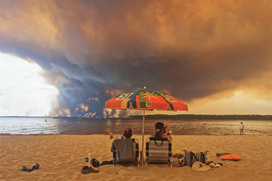 Two people relaxing on a beach under a colorful umbrella as a wildfire burns across a lake in the background.