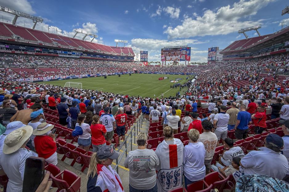 A stadium full of people watching an England vs. New Zealand soccer match on a sunny day.