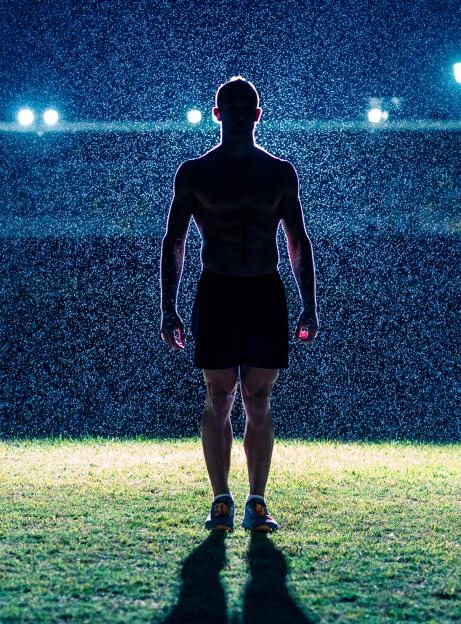 Silhouette of a muscular male athlete standing on a stadium field in the rain at night, illuminated by floodlights.