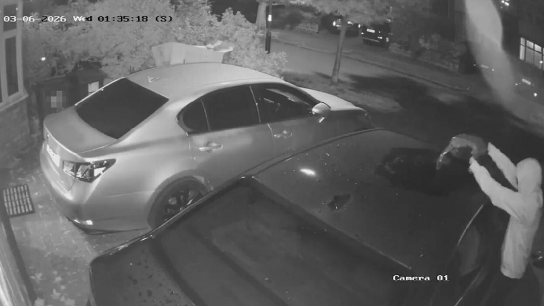 Security camera footage of an individual in a white hoodie tampering with a car.