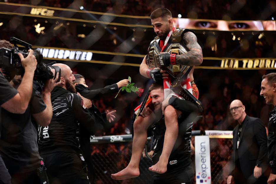 UFC fighter Ilia Topuria, with championship belts, is carried on another man's shoulders while holding a red rose.