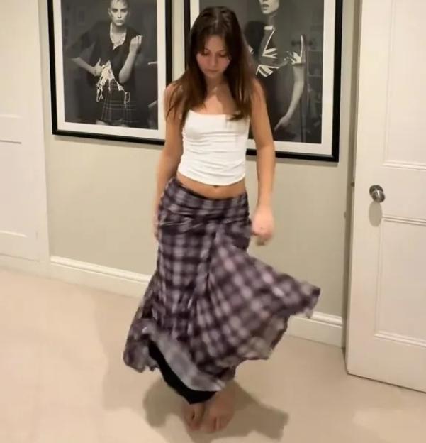A woman in a white tank top and a flowing, long plaid skirt dancing, with two framed black and white portraits on the wall behind her.