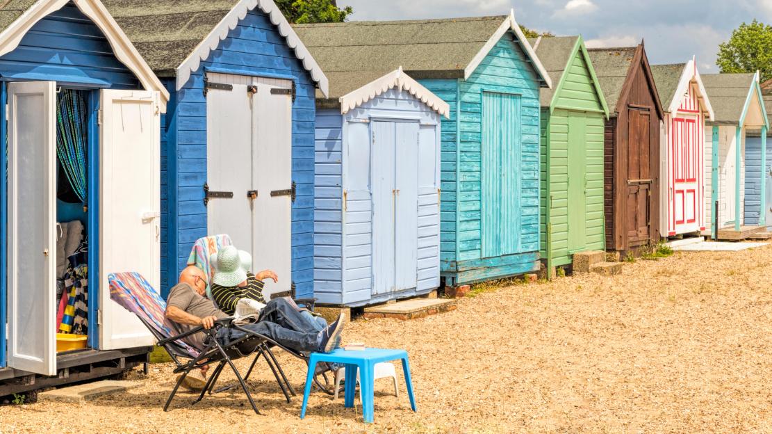 Couple relaxing in chairs in front of a row of brightly colored beach huts on a sunny day.