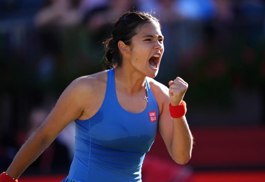 A female tennis player in a blue top and red wristband celebrates with a clenched fist and open mouth.