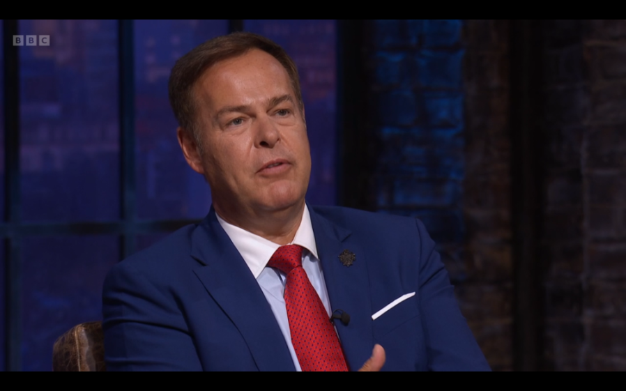 A man in a blue suit and red tie speaking in front of a dark, brick-walled background.