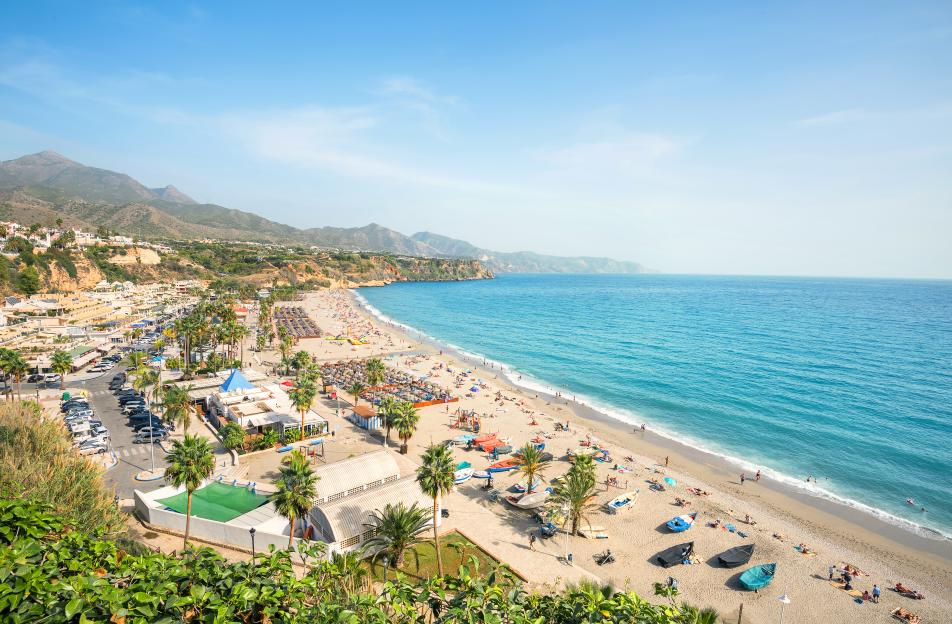 Overhead view of Nerja Beach in Malaga, Spain, showing the sandy shore filled with people, colorful umbrellas, and boats, with a town nestled against mountains in the background.