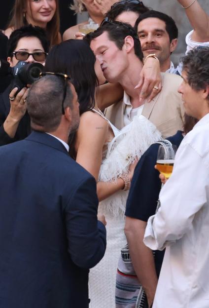 Dua Lipa and Callum Turner kissing at a party while guests watch and take photos.