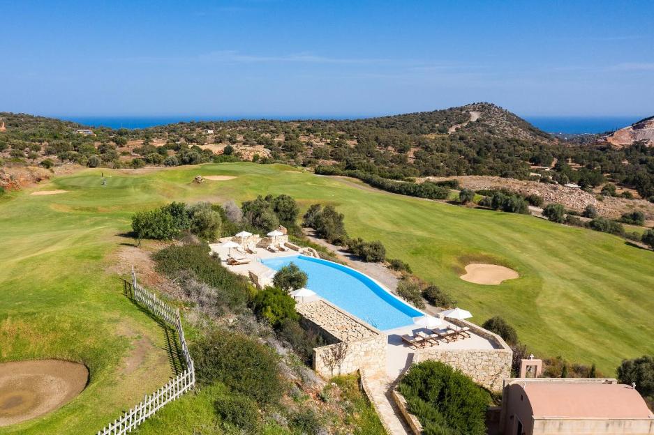 An aerial view of the Crete Golf Club in Hersonissos, with a bright blue swimming pool and lounge chairs overlooking a green golf course, surrounded by rolling hills and the ocean in the distance.