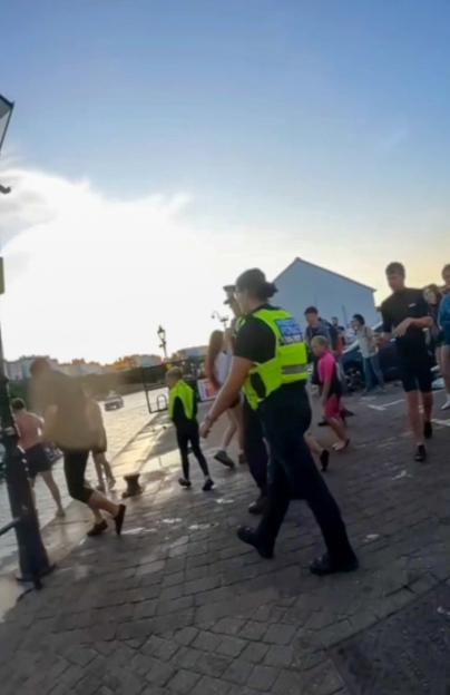 A female police officer in a high-visibility vest walks along a cobblestone path next to a body of water, with other people around.