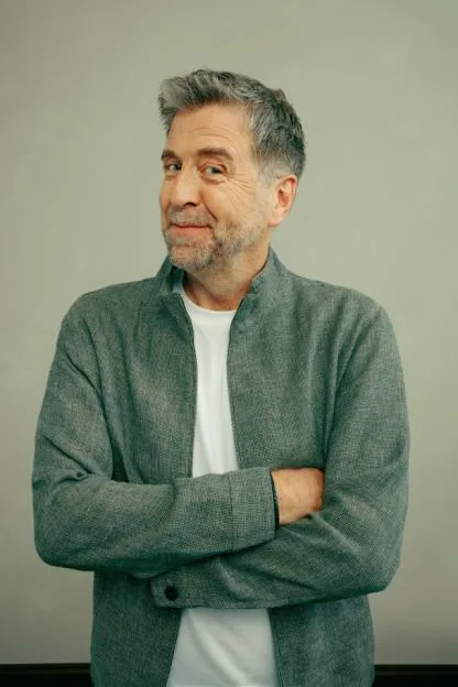 Portrait of a middle-aged man with graying hair and a beard, wearing a grey jacket and white t-shirt, standing with arms crossed against a plain background.