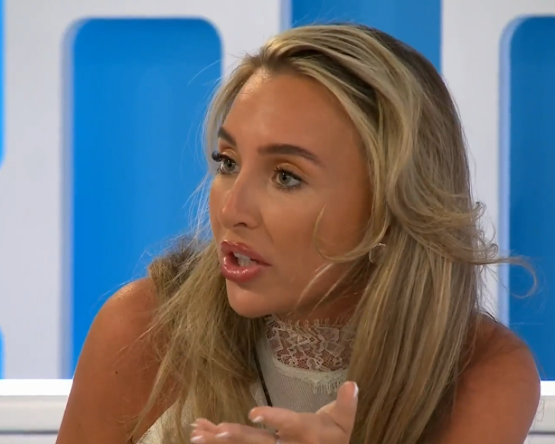 A woman with long blonde hair and a white lace top talking with her hand raised.