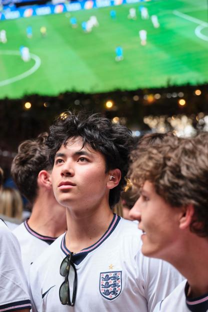 A man wearing a white England football jersey with "CheerPlugs" in his ear, watching a football game on a large screen.