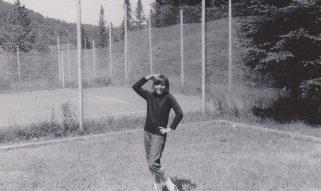A young person stands on a grassy area, smiling at the camera, with a tennis court and forested hills behind them.