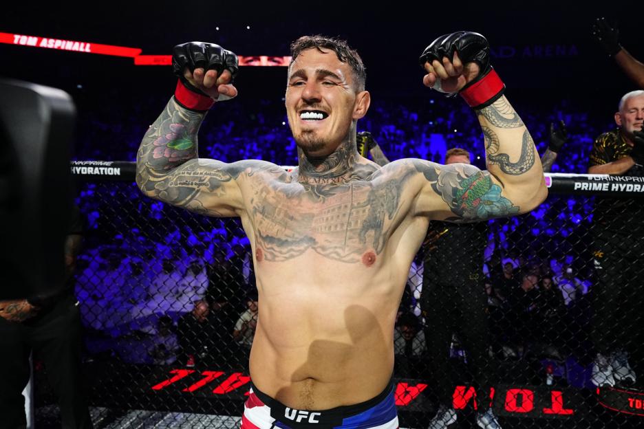 UFC fighter Tom Aspinall, shirtless and tattooed, flexing his biceps and smiling with a mouthguard.