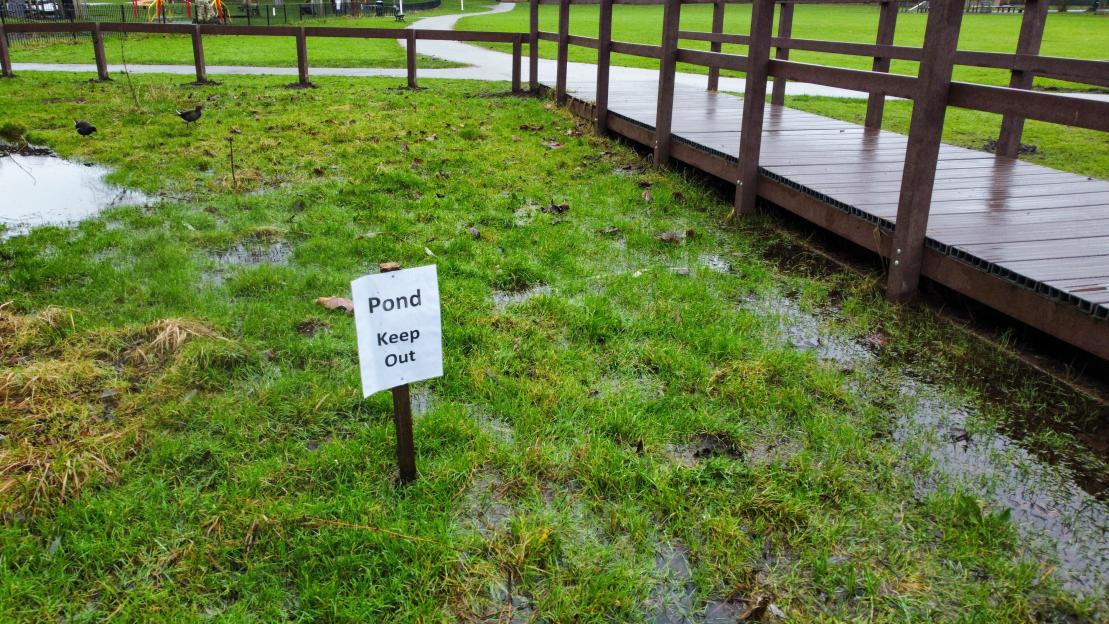A sign reading "Pond Keep Out" placed in a shallow puddle of water next to a fenced walkway in a park.