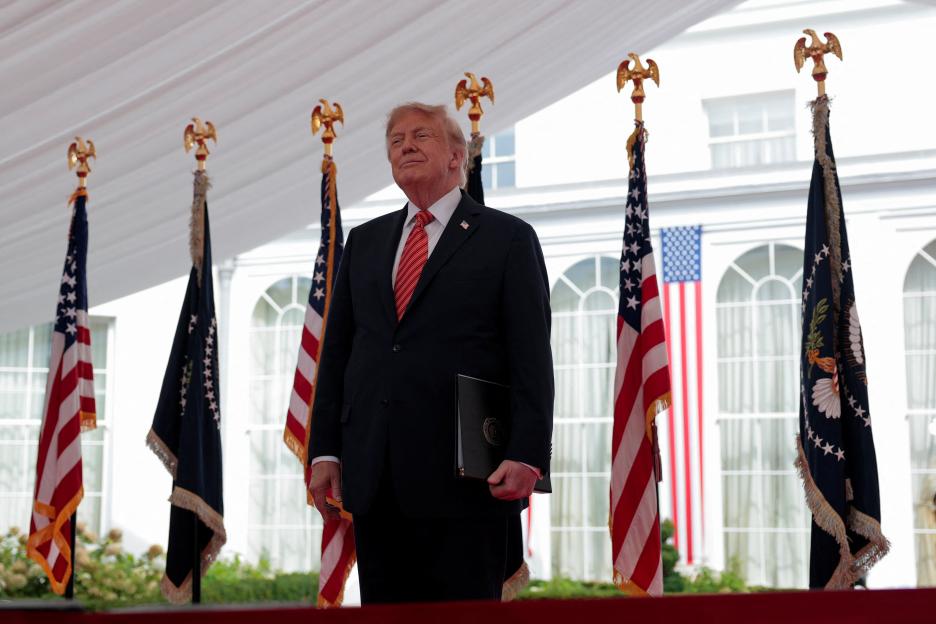 U.S. President Donald Trump stands holding a folder, flanked by American and presidential flags.