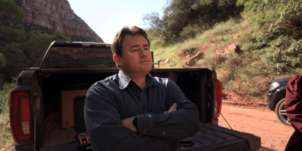Samuel Bateman standing in front of a pickup truck with arms crossed in Trust Me: The False Prophet.