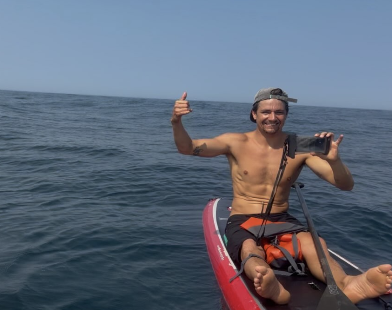 A man on a paddleboard in the ocean, giving a thumbs-up and holding a phone.