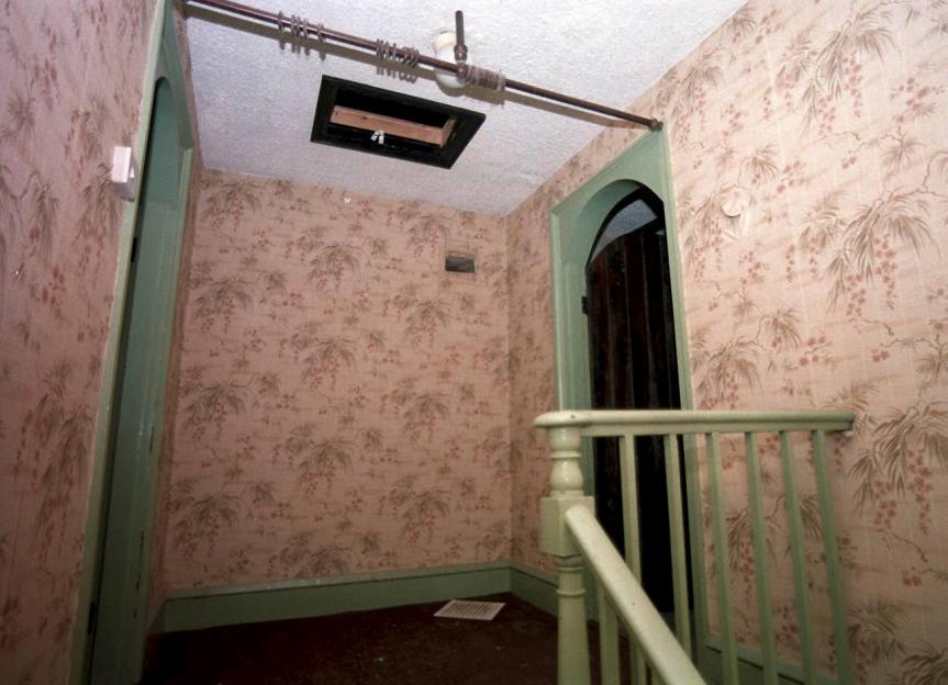 Hallway inside 25 Cromwell Street, the home of Fred and Rose West, with floral wallpaper, a light green banister, and an open doorway.
