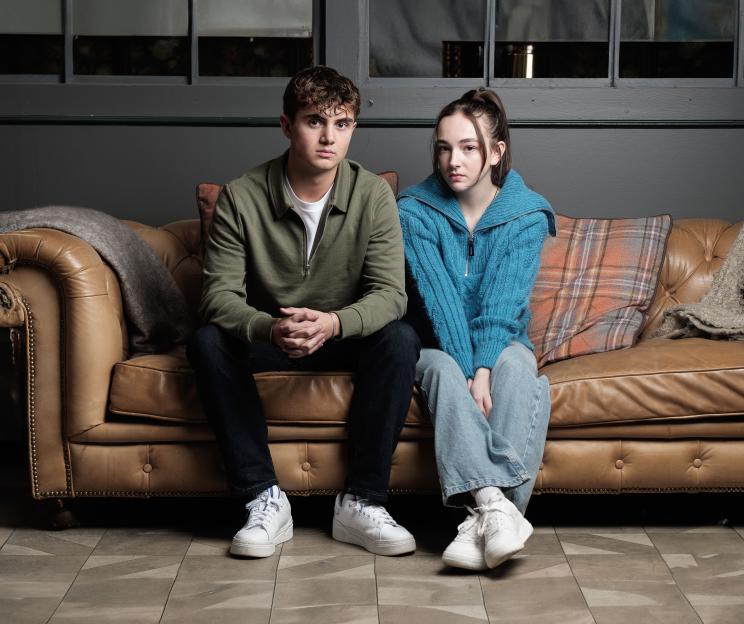 A young man and woman sit on a brown leather couch looking straight ahead.