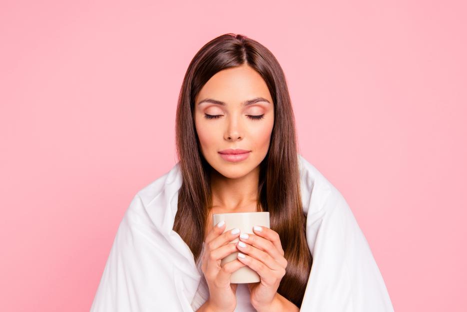 Young woman wrapped in a white blanket, holding a mug of coffee with closed eyes, against a pink background.