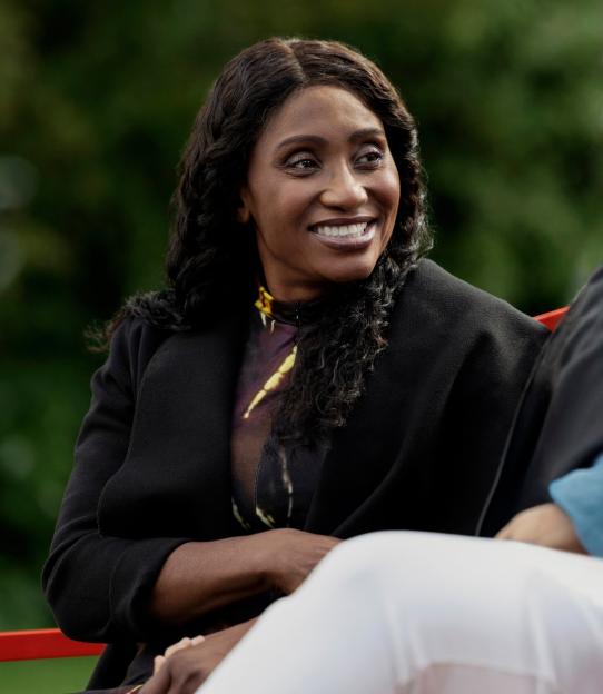 A woman with dark curly hair, wearing a black coat over a patterned shirt, smiles and looks to her right.
