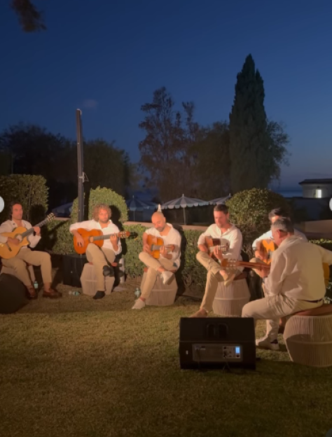 Five men in white and beige clothing sitting in a circle on a lawn playing classical guitars at night.