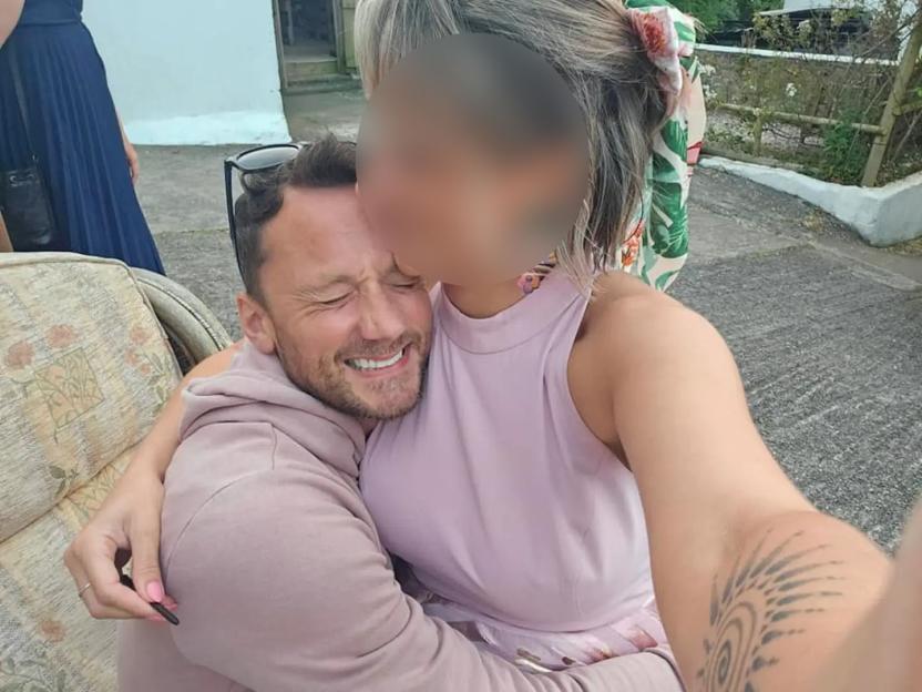 A man with his eyes closed, smiling widely, is hugged by a woman whose face is obscured.