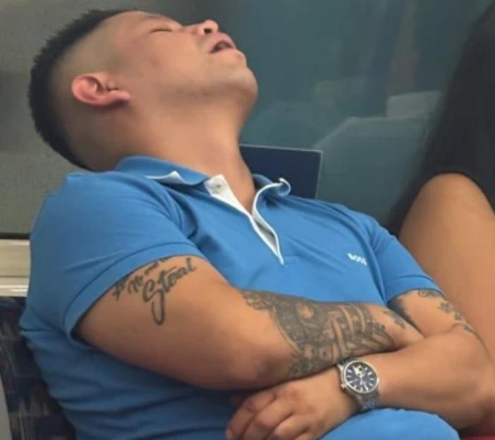 A man in a blue polo shirt and watch with tattoos on his arms, sleeping with his mouth open.