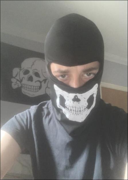 Alfie Coleman wearing a black balaclava with a white skull design over his mouth and nose, with a flag depicting a skull in the background.