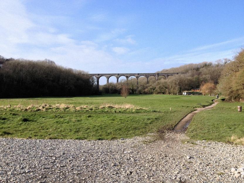 A long stone viaduct arches over a green, grassy field with a walking path, bordered by a forest.