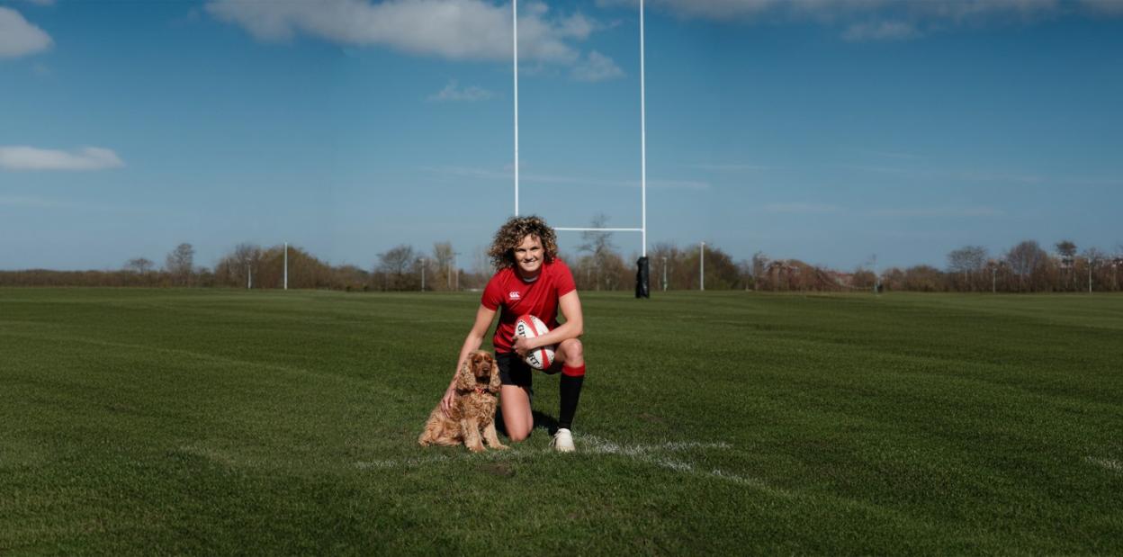 A female rugby player in a red shirt with a cocker spaniel on a green field.