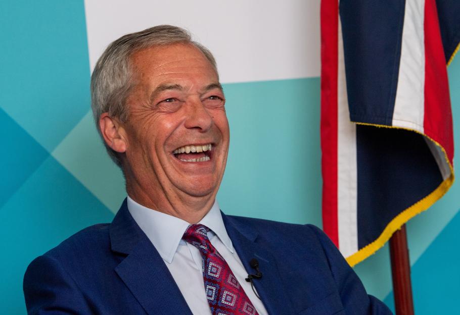 Nigel Farage laughing heartily with a Union Jack flag in the background.